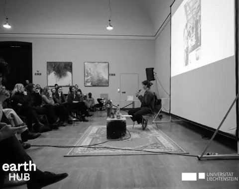 Schwarz-Weiß-Foto eines Vortrags an der Universität Liechtenstein. Eine Person sitzt vor einem Publikum und spricht, während eine Präsentation auf eine Leinwand projiziert wird. Im Vordergrund sind Logos von "earth HUB" und "Universität Liechtenstein" sichtbar.