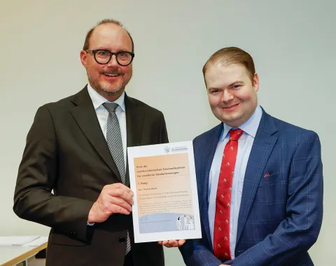 Zwei Männer in Anzügen halten gemeinsam ein Zertifikat in die Kamera. Der Hintergrund ist neutral. Sie lächeln und stehen in einem Raum, der für eine feierliche Veranstaltung geeignet wirkt.