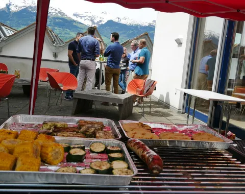 Alumni-Treffen an der Universität Liechtenstein: Eine Gruppe von Menschen steht auf einer Terrasse und unterhält sich bei einem BBQ. Im Vordergrund sind gegrillte Speisen zu sehen. Im Hintergrund sind Berge unter einem blauen Himmel sichtbar.