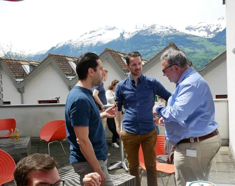 Alumni-Treffen an der Universität Liechtenstein: Ehemalige Studierende unterhalten sich auf einer sonnigen Terrasse mit Bergblick im Hintergrund.