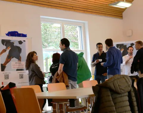 Eine Gruppe von Menschen steht in einem Raum der Universität Liechtenstein und unterhält sich. Im Hintergrund sind Poster und ein Fenster zu sehen. Die Atmosphäre wirkt entspannt und gesellig.