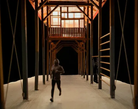 Ein Modell eines architektonischen Entwurfs zeigt eine überdachte Struktur mit sichtbaren Holzbalken. Eine Person läuft auf einem Weg unter der Konstruktion. Das Design kombiniert traditionelle Bauweise mit modernen Elementen und betont lineare Formen.