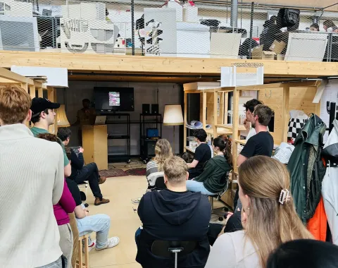 Eine Gruppe von Studierenden sitzt in einem Architekturatelier und hört einem Vortrag zu. Im Hintergrund sind Regale und Arbeitsmaterialien zu sehen.