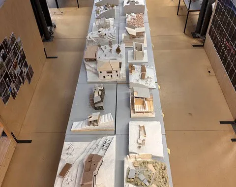 Eine Reihe von Architekturmodellen auf Tischen in einem Studio. Die Modelle zeigen verschiedene Gebäude und Landschaften, die im Rahmen eines Projekts des Advanced Studios Upcycling der Universität Liechtenstein erstellt wurden. Im Hintergrund sind Regale und weitere Arbeitsbereiche zu sehen.