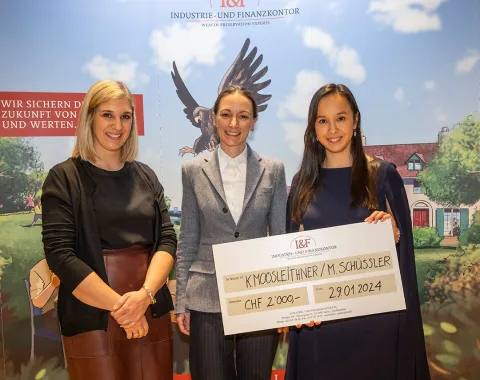 Drei Personen stehen lächelnd vor einem Hintergrund mit dem Logo der Industrie- und Finanzkontor. Eine Person hält einen großen Scheck mit der Aufschrift "CHF 2,000" und einem Datum. Im Hintergrund ist ein stilisiertes Bild mit einem Adler und einem Haus zu sehen.