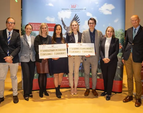 Gruppe von acht Personen posiert mit zwei großen Schecks bei der Verleihung des I&F Family Wealth Preservation Awards an der Universität Liechtenstein. Die Studierenden wurden für ihre herausragenden Strukturierungsvorschläge ausgezeichnet. Im Hintergrund ist ein Banner des Industrie- und Finanzkontors zu sehen.