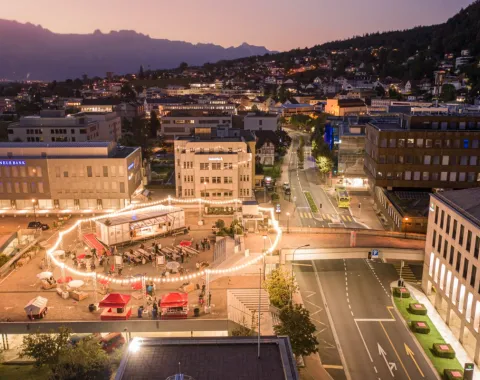 Luftaufnahme einer städtischen Umgebung bei Dämmerung. Im Zentrum des Bildes ist ein beleuchteter Platz mit einem kreisförmigen Lichterkranz zu sehen, umgeben von Gebäuden. Menschen versammeln sich um das Base Camp, eine modulare Plattform. Im Hintergrund erstreckt sich eine hügelige Landschaft.