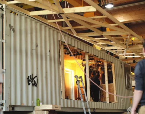 Ein großer, umgebauter Container in einer Werkhalle, mit sichtbaren Holzverstärkungen und einer Leiter, symbolisiert die flexible Nutzung des Base Camps der Universität Liechtenstein für Stadtentwicklungsprojekte.