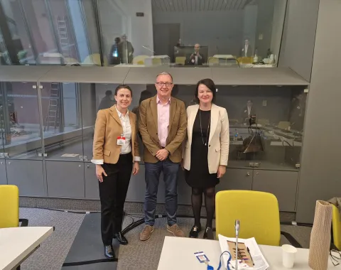 Drei Personen stehen in einem modernen Konferenzraum des Europarats in Straßburg. Sie lächeln in die Kamera. Im Hintergrund sind weitere Personen und Konferenztische zu sehen.