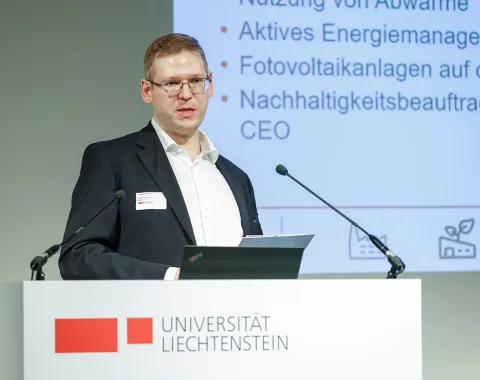 Ein Mann hält einen Vortrag an einem Rednerpult der Universität Liechtenstein. Im Hintergrund ist eine Präsentation mit Themen wie Energiemanagement und Photovoltaikanlagen zu sehen.