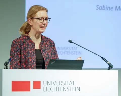 Eine Person hält eine Rede an einem Rednerpult mit dem Logo der Universität Liechtenstein. Im Hintergrund ist eine Präsentation zu sehen.