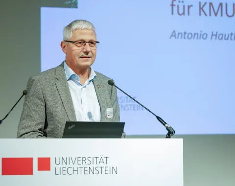 Ein Redner steht an einem Podium mit dem Logo der Universität Liechtenstein und spricht in ein Mikrofon. Im Hintergrund ist eine Präsentationsfolie zu sehen.