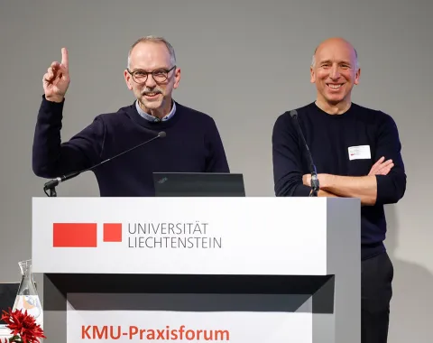 Zwei Männer stehen lächelnd hinter einem Rednerpult mit dem Logo der Universität Liechtenstein. Einer hebt die Hand, während der andere die Arme verschränkt. Im Vordergrund sind rote Blumen zu sehen.