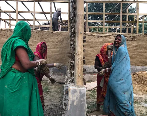 Frauen in bunten Saris arbeiten gemeinsam an einem Lehmgebäude. Im Hintergrund ist eine Holzkonstruktion zu sehen. Sie lächeln und sind in den Bauprozess vertieft.