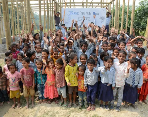Eine große Gruppe fröhlicher Kinder steht vor einem im Bau befindlichen Schulgebäude in Bahuarwa, Indien. Im Hintergrund halten zwei Erwachsene ein Schild mit der Aufschrift "Thank You Very Much". Die Kinder lächeln und winken in die Kamera.