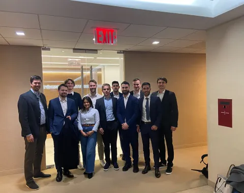 Eine Gruppe von Studierenden in Business-Kleidung steht in einem modernen Bürogebäude. Im Hintergrund ist eine Glastür zu sehen. Die Szene wirkt professionell und vermittelt den Eindruck einer Exkursion oder eines geschäftlichen Treffens.