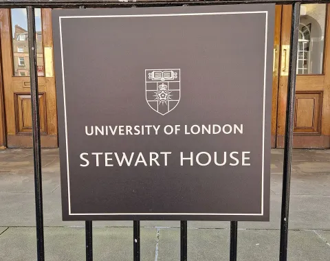 Ein Schild vor dem Eingang eines Gebäudes mit der Aufschrift "University of London, Stewart House". Im Hintergrund sind Holztüren und die Hausnummer 32 zu sehen.