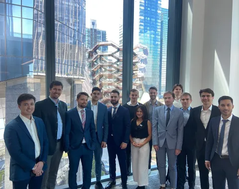 Eine Gruppe von Studierenden in Business-Kleidung steht vor großen Fenstern mit Blick auf moderne Architektur und Wolkenkratzer in New York City.