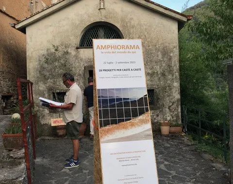 Ein Mann steht vor einem historischen Gebäude in Castè, Italien, und betrachtet ein Plakat mit der Aufschrift "Amphiorama". Das Plakat informiert über ein Projekt der Universität Liechtenstein im Rahmen des Advanced Studios Upcycling. Im Hintergrund sind die malerischen Hügel der Cinque Terre zu sehen.
