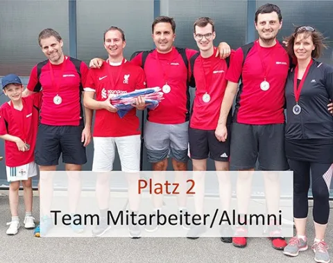 Gruppe von sechs Personen in roten Trikots mit Medaillen, die das Team "Mitarbeiter/Alumni" repräsentieren, posiert für ein Foto. Ein Kind steht links daneben. Ein Schild zeigt "Platz 2". Hintergrund: Gebäude mit Glasfront.