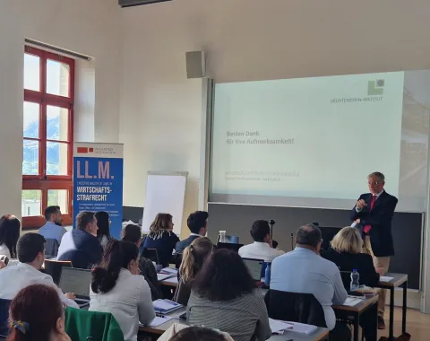 Ein Vortragender steht vor einer Gruppe von Teilnehmenden in einem Seminarraum der Universität Liechtenstein. Auf der Leinwand ist eine Präsentationsfolie mit dem Text "Besten Dank für Ihre Aufmerksamkeit!" zu sehen. Im Hintergrund ist ein Banner mit der Aufschrift "LL.M. Wirtschafts-Strafrecht" sichtbar.