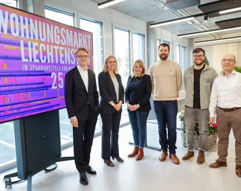 Gruppe von sechs Personen steht vor einem Bildschirm mit der Aufschrift "Wohnungsmarkt Liechtenstein". Die Personen lächeln und sind in einem modernen Raum mit großen Fenstern.