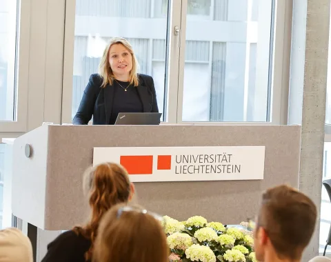 Eine Person steht an einem Rednerpult der Universität Liechtenstein und spricht zu einem Publikum. Im Hintergrund sind große Fenster zu sehen. Vor dem Pult sind Blumen arrangiert.