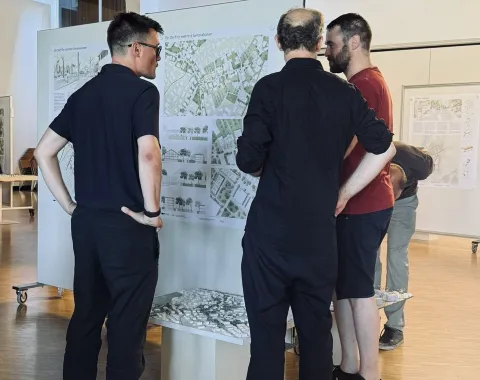 Drei Personen betrachten Architekturpläne und ein Modell in einem Ausstellungsraum der Universität Liechtenstein. Die Szene zeigt eine Diskussion über ortsbauliche Strategien zur Entwicklung des Zentrums von Ruggell.