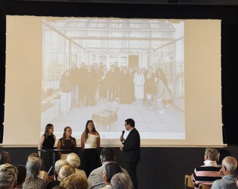 Eine Gruppe von Studierenden präsentiert ihre Projekte zur Ortsentwicklung in Ruggell vor einem Publikum. Im Hintergrund ist ein Bild der Studierenden auf einer Leinwand zu sehen.