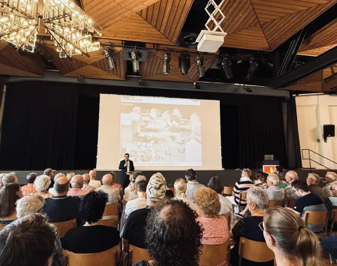 Eine Person hält einen Vortrag vor einem Publikum in einem Saal. Auf der Bühne ist eine große Leinwand mit einer Präsentation zu sehen. Der Raum hat eine moderne Holzdecke mit Beleuchtung.
