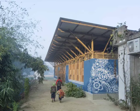 Kinder gehen auf einem sandigen Weg an einem blau bemalten Bambusgebäude mit weißem Ornamentmuster vorbei.