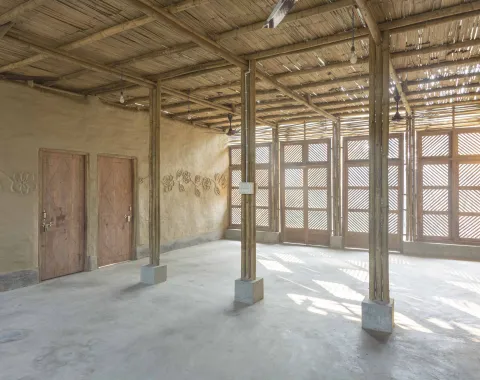 Ein sonnendurchfluteter Raum aus Bambus und Lehm mit zwei Holztüren und dekorativen Blumenreliefs an der Wand.