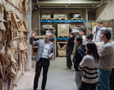 Mann zeigt auf eine Wand mit Holzvorlagen und erklärt sie einer Gruppe von Studierenden 