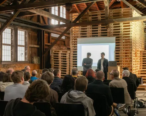 Zwei Männer stehen in einem Dachgeschoss udn präsentieren ein Projekt vor einem sitzenden Publikum