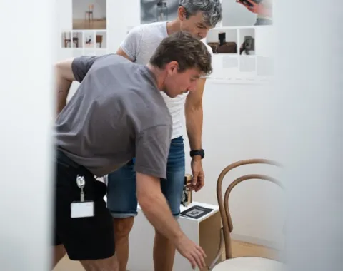 Zwei Männer bücken sich zu einem Kaffeehausstuhl in einer Ausstellung