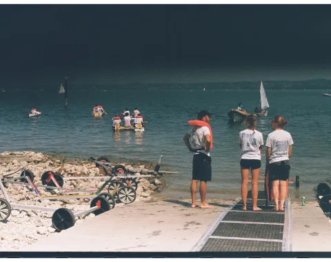 Mehrere Menschen stehen vor einem See und schauen auf Boote. 