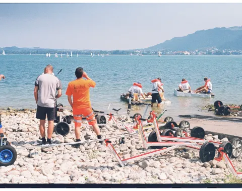 Mehrere Menschen am Ufer schauen auf selbstgebaute Boote in einem See. 