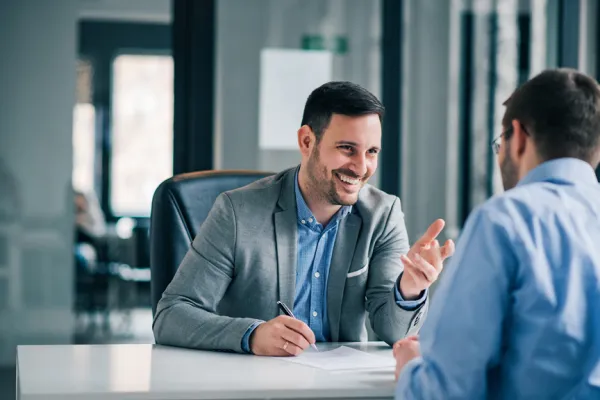 Ein lächelnder Mann in einem grauen Blazer sitzt an einem Schreibtisch und spricht gestikulierend mit einer anderen Person, die ihm gegenüber sitzt. Die Szene vermittelt eine Atmosphäre des professionellen Austauschs und der Beratung.
