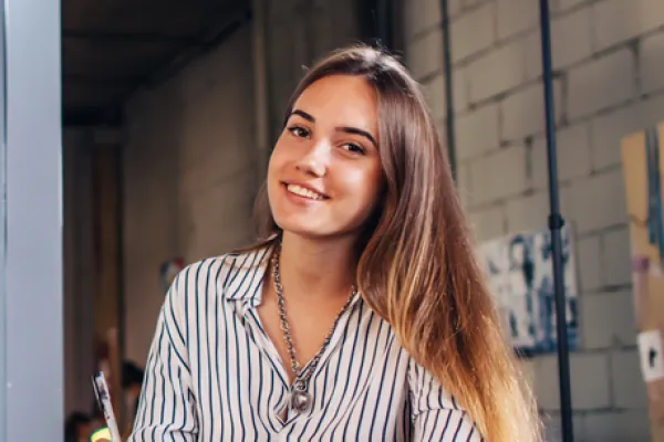 Eine junge Frau sitzt an einem Schreibtisch mit einem Notizbuch und lächelt freundlich in die Kamera. Sie trägt ein gestreiftes Hemd, und im Hintergrund ist eine kreative Büroumgebung zu sehen. Der Schreibtisch ist mit Laptop, Notizen und einer Kaffeetasse bedeckt, während sie einen Stift in der Hand hält, bereit zu schreiben.