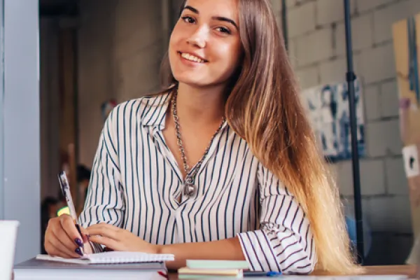 Eine junge Frau sitzt an einem Schreibtisch mit einem Notizbuch und lächelt freundlich in die Kamera. Sie trägt ein gestreiftes Hemd, und im Hintergrund ist eine kreative Büroumgebung zu sehen. Der Schreibtisch ist mit Laptop, Notizen und einer Kaffeetasse bedeckt, während sie einen Stift in der Hand hält, bereit zu schreiben.