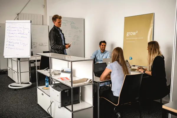Vier Studierende arbeiten im Team an einem Projekttisch, mit Whiteboard und Flipchart im Hintergrund