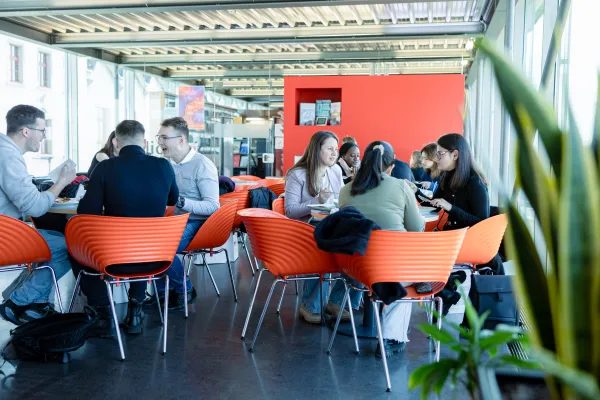 Lebendige Szene in der Universitätscafeteria mit mehreren Gruppen Studierender beim gemeinsamen Mittagessen und Gesprächen.