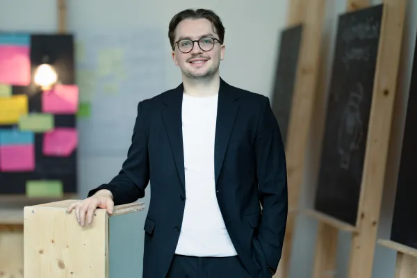 Student der Universität Liechtenstein im schwarzen Jacket lächelt in die Kamera – Campusaufnahme in freundlicher Lernumgebung.