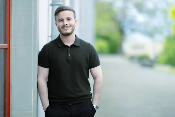 Ein männlicher Studierender im Masterstudiengang Information Systems in einem dunkelgrünen Polohemd steht locker an eine Wand gelehnt, mit den Händen in den Hosentaschen und einem dezenten Lächeln.