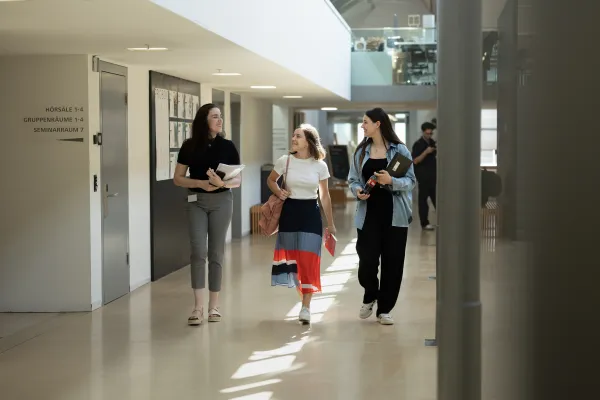 Drei Studentinnen gehen lächelnd durch das lichtdurchflutete Foyer mit Unterlagen in der Hand.