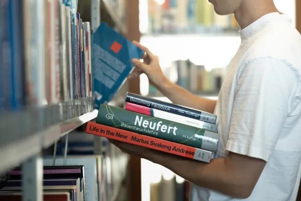 Student wählt Fachliteratur aus dem Bücherregal der Universitätsbibliothek Liechtenstein, mehrere Bücher im Arm.
