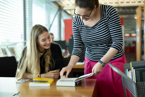 Eine Bibliotheksmitarbeiterin zeigt einer lächelnden Studentin Inhalte aus einem geöffneten Buch.