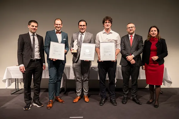 Eine Gruppe von sechs Personen steht nebeneinander auf einer Bühne. Drei Personen in der Mitte halten Auszeichnungen und Urkunden. Alle sind formell gekleidet und lächeln in die Kamera. Im Hintergrund sind weiße Tischdecken sichtbar.