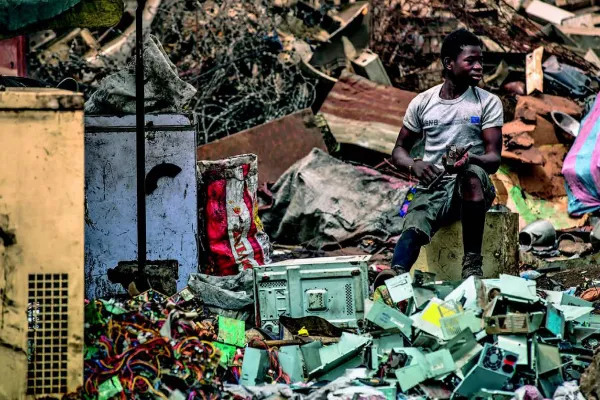 Ein Mann sitzt auf einem Hocker inmitten von Elektroschrott auf einer Müllhalde. Um ihn herum sind alte Geräte und Kabel zu sehen. Die Szene zeigt die Problematik von Elektroschrott und dessen Auswirkungen auf die Umwelt.