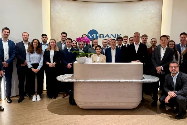 Gruppe von Studierenden und Fachleuten in formeller Kleidung steht in der Lobby der VP Bank. Im Hintergrund ist das Logo der Bank sichtbar. Die Szene vermittelt eine professionelle und lernorientierte Atmosphäre.